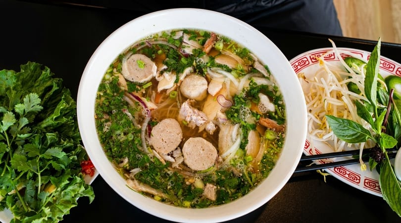 Overhead view of a large bowl of pho with meat, herbs, and a side of fresh garnishes.