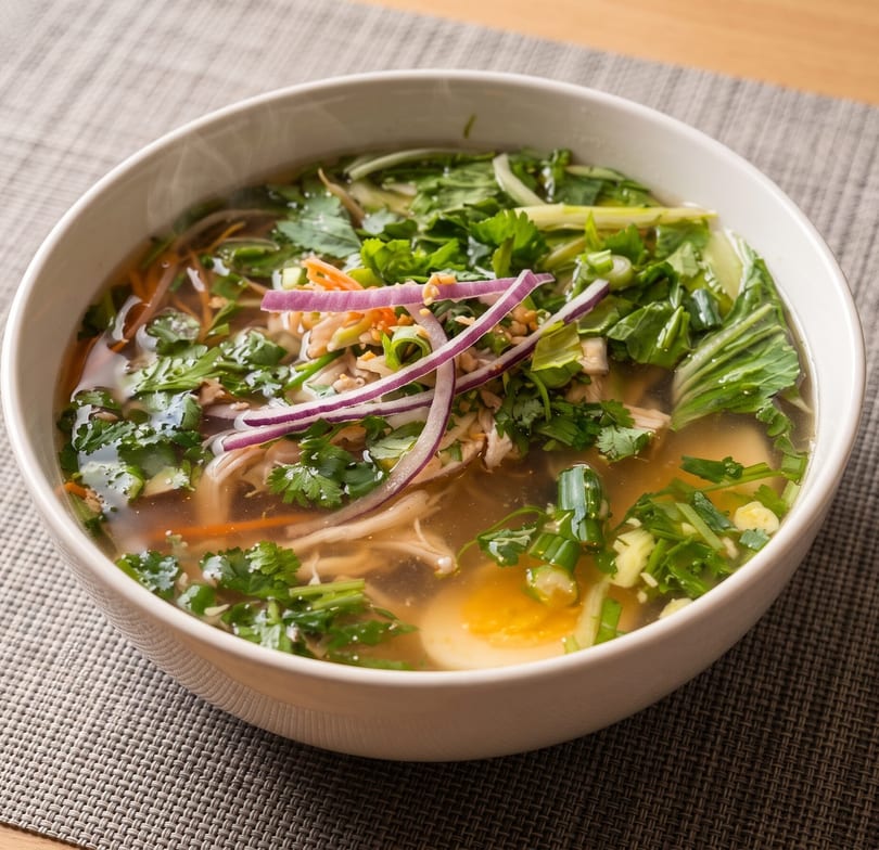 A steaming bowl of pho with chicken, herbs, red onions, and a soft-boiled egg.