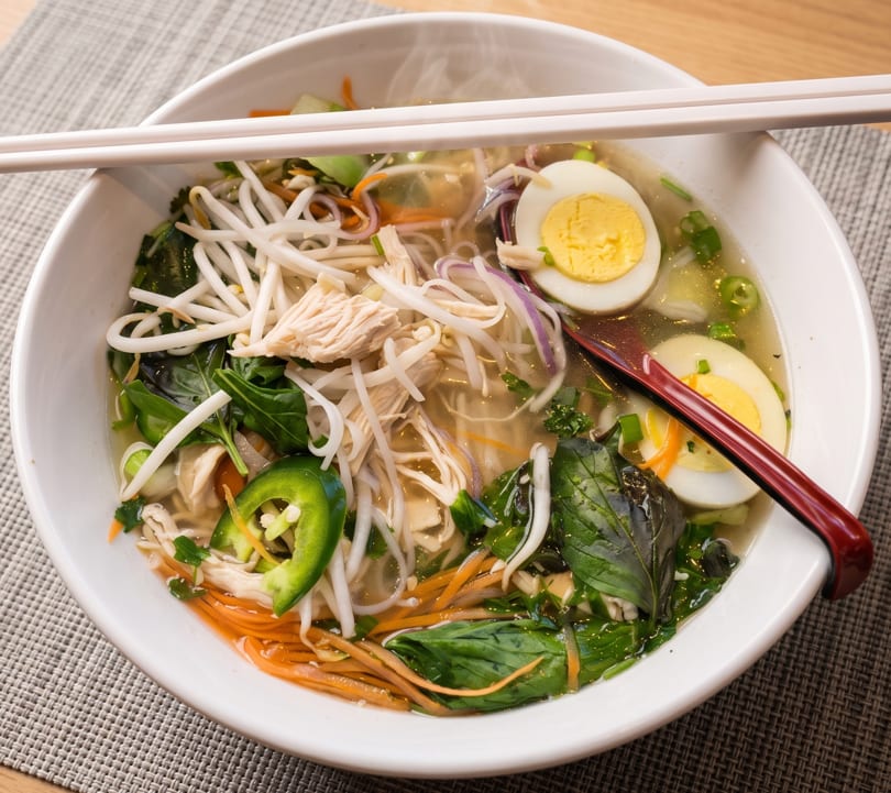 Steaming bowl of pho with chicken, bean sprouts, herbs, jalapenos, and hard-boiled eggs.